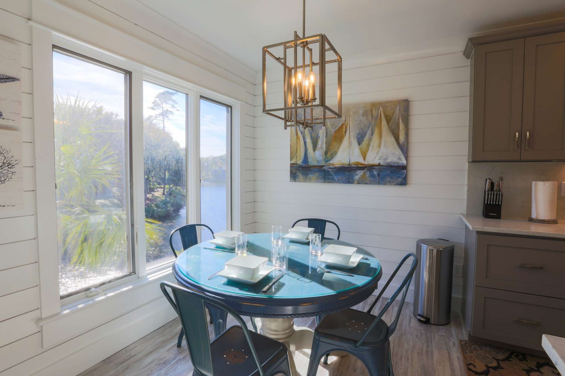 A dining area with a round glass table, metal chairs, a hanging light fixture, and a painting of sailboats.