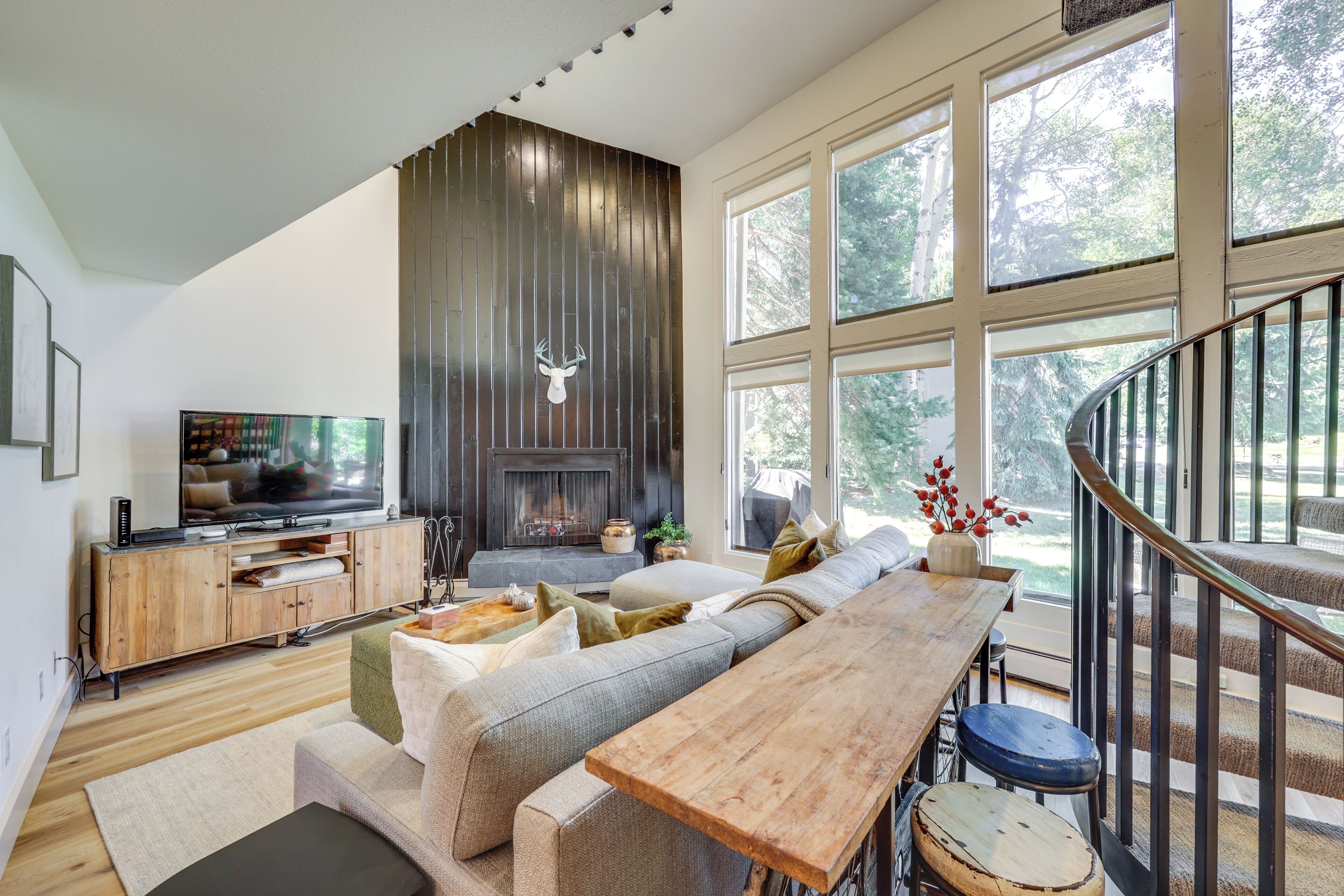 A modern living room with a fireplace, large windows, a wooden bench, and a staircase.