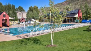 Piscina com espreguiçadeiras, uma pequena árvore e edifícios ao fundo.