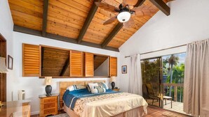 Un dormitorio con techo de madera, una cama grande con colcha azul y blanca, una mesita de noche con lámpara y vistas a un balcón con cortinas.