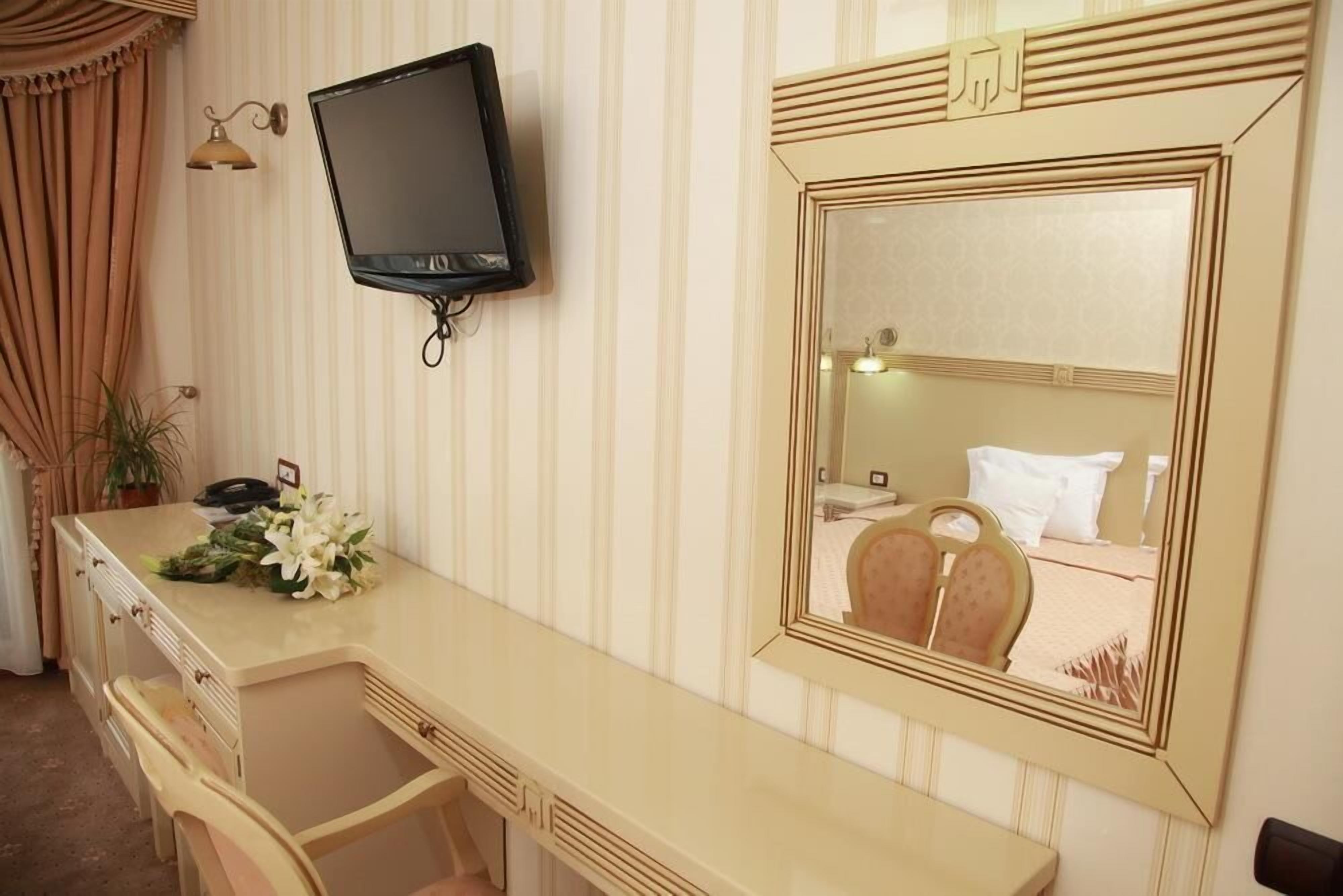 A hotel room with a long, beige vanity, a flat-screen TV mounted on the wall, and a large mirror with a decorative frame.