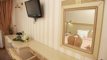 A hotel room with a long, beige vanity, a flat-screen TV mounted on the wall, and a large mirror with a decorative frame.