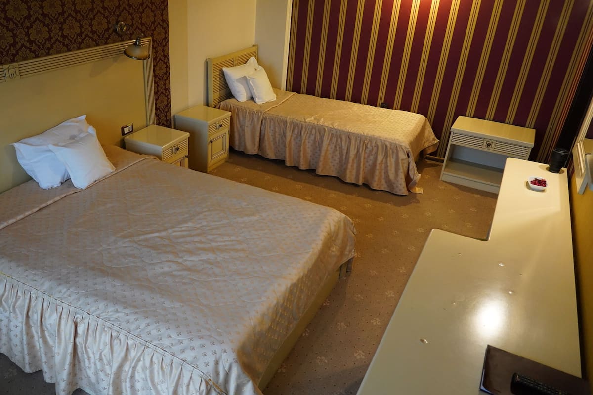 A hotel room with two beds, a bedside table, a lamp, a heater, and a small table with a bowl of fruit.
