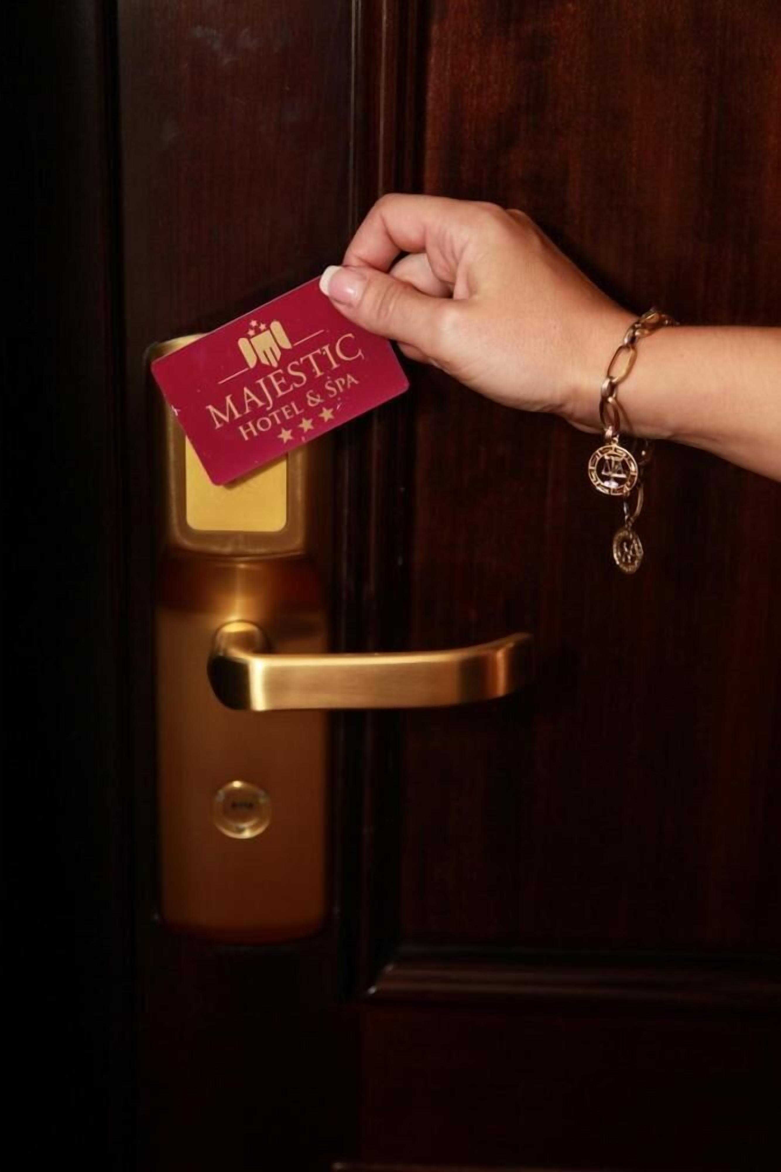 Eine Hand hält eine rote Karte mit dem Text 'MAJESTIC HOTEL & SPA', vor einer Tür mit Messinggriffen.