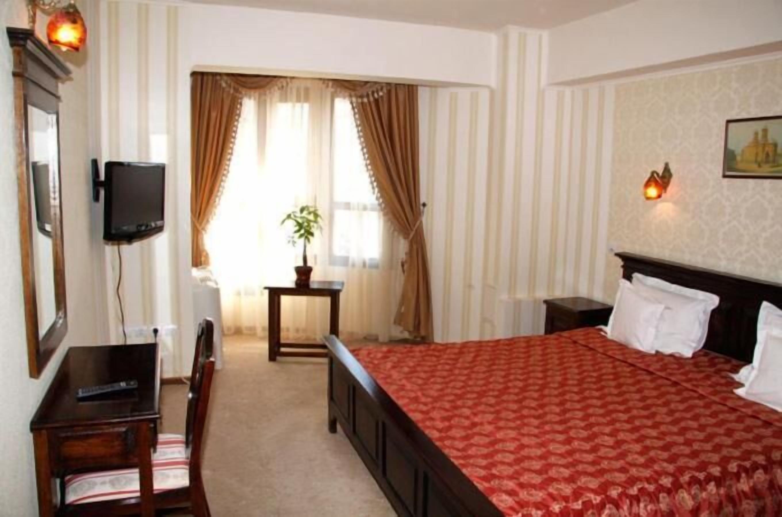 A hotel room with a bed, a desk, a chair, a TV, a small table with a plant, and a window with curtains.