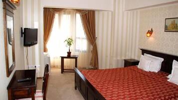 A hotel room with a bed, a desk, a chair, a TV, a small table with a plant, and a window with curtains.