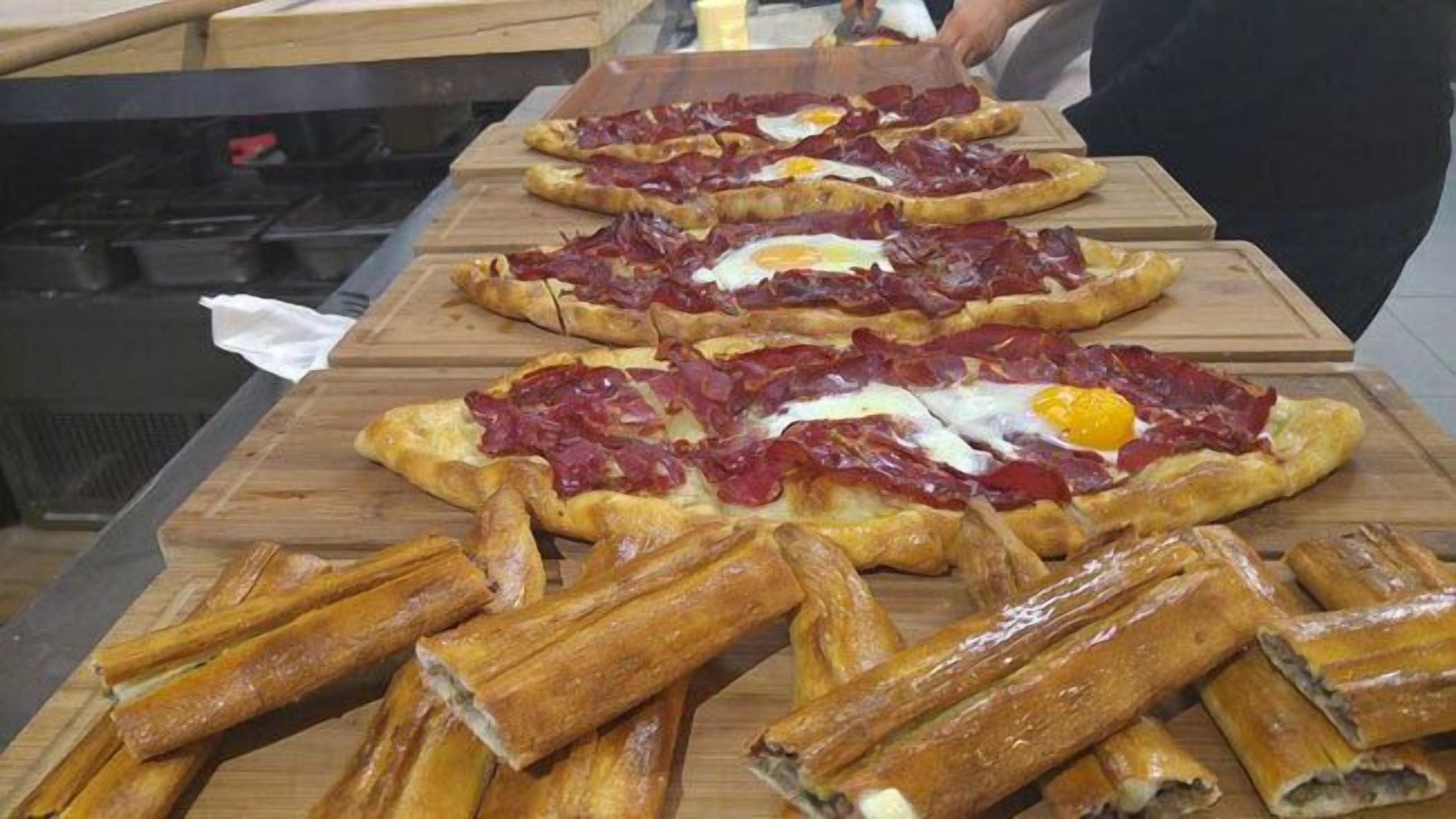 Een keukenblad met meerdere pizza's belegd met vlees en eieren, en een stapel stokbroden op de voorgrond.