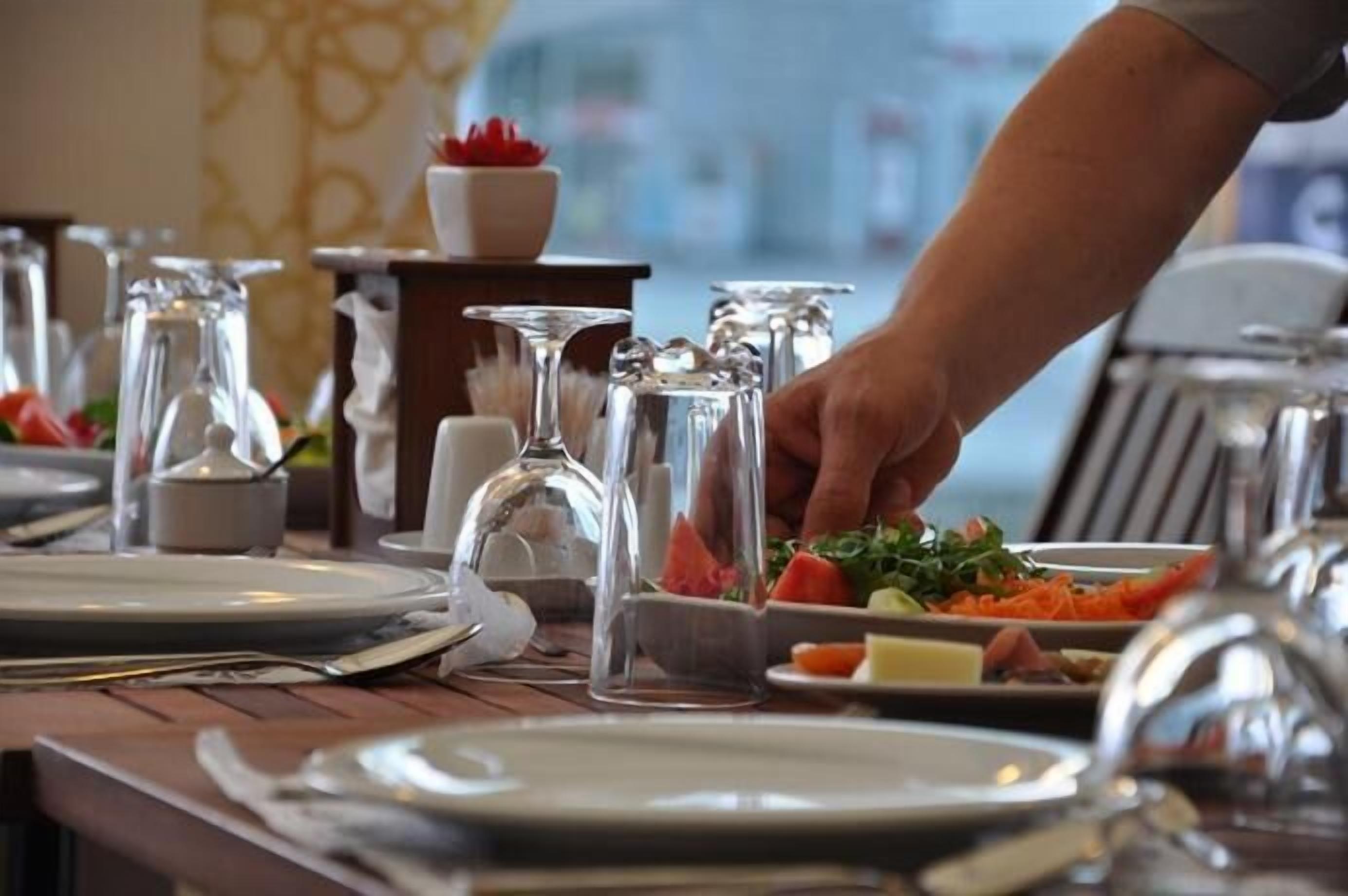 Una mesa de comedor dispuesta con platos, cubiertos y cristalería, con la mano de una persona alcanzando un plato.
