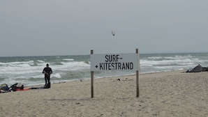 A beach with a sign reading "SURF-KITESTRAND" and a person standing near a kiteboard.