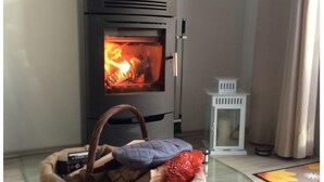A modern wood-burning stove with a glass door, a wicker basket with blankets and a pillow on the floor, and a small white table beside it.