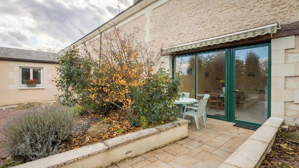Un patio con mesa y sillas, un pequeño jardín con diversas plantas y un ventanal corredizo que da a un espacio interior.