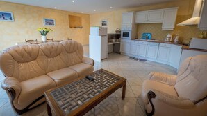 Una sala de estar con un sofá color beige, una mesa de centro de vidrio y una zona de cocina con armarios blancos y salpicadero de azulejos.