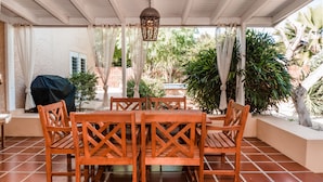 A covered patio with a wooden table and chairs, surrounded by plants and a grill. - A private vacation villa with gorgeous pool. (Kralendijk)