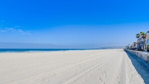 A wide sandy beach with footprints, a clear blue sky, and a row of palm trees along the edge.