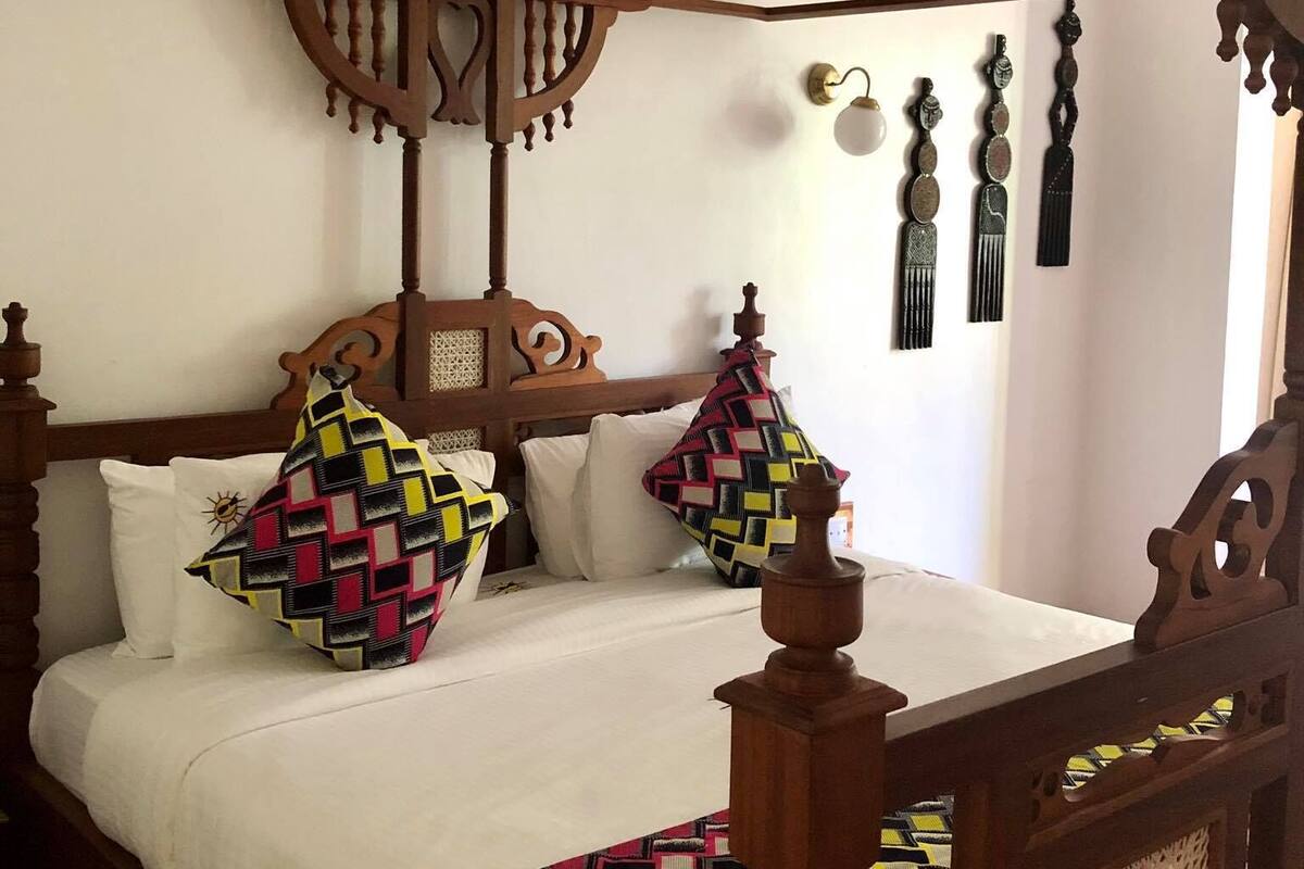 Uma cama de dossel com estrutura de madeira e cortina, roupa de cama branca e almofadas estampadas e coloridas.