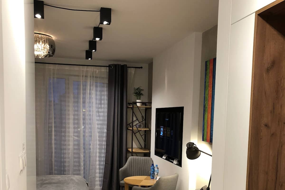 Quarto de hotel moderno com cama, escrivaninha, cadeira, mesinha, televisão e planta.