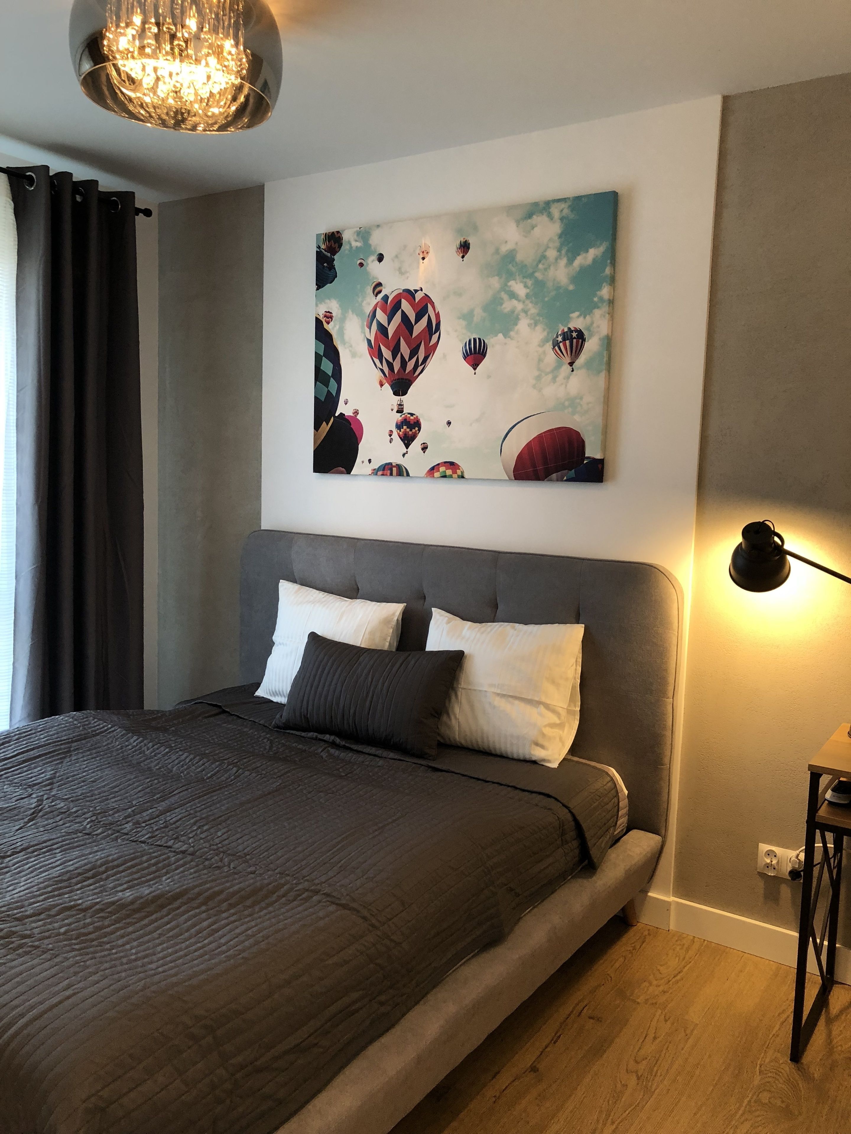 Une chambre moderne avec un grand lit, un lustre, un tableau représentant des montgolfières et une table de chevet avec une lampe.
