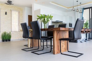 A modern dining area with a wooden table and black chairs, a vase with green plants, and a kitchen in the background.