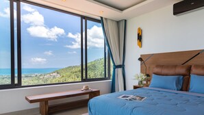 A bedroom with a large window, a wooden bench, a bed with blue bedding, and a nightstand with a lamp. - Villa Victoria (Koh Samui)