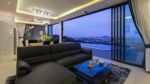 A modern living room with a large sectional sofa, a glass coffee table, and a view of a cityscape at dusk. - Villa Victoria (Koh Samui)