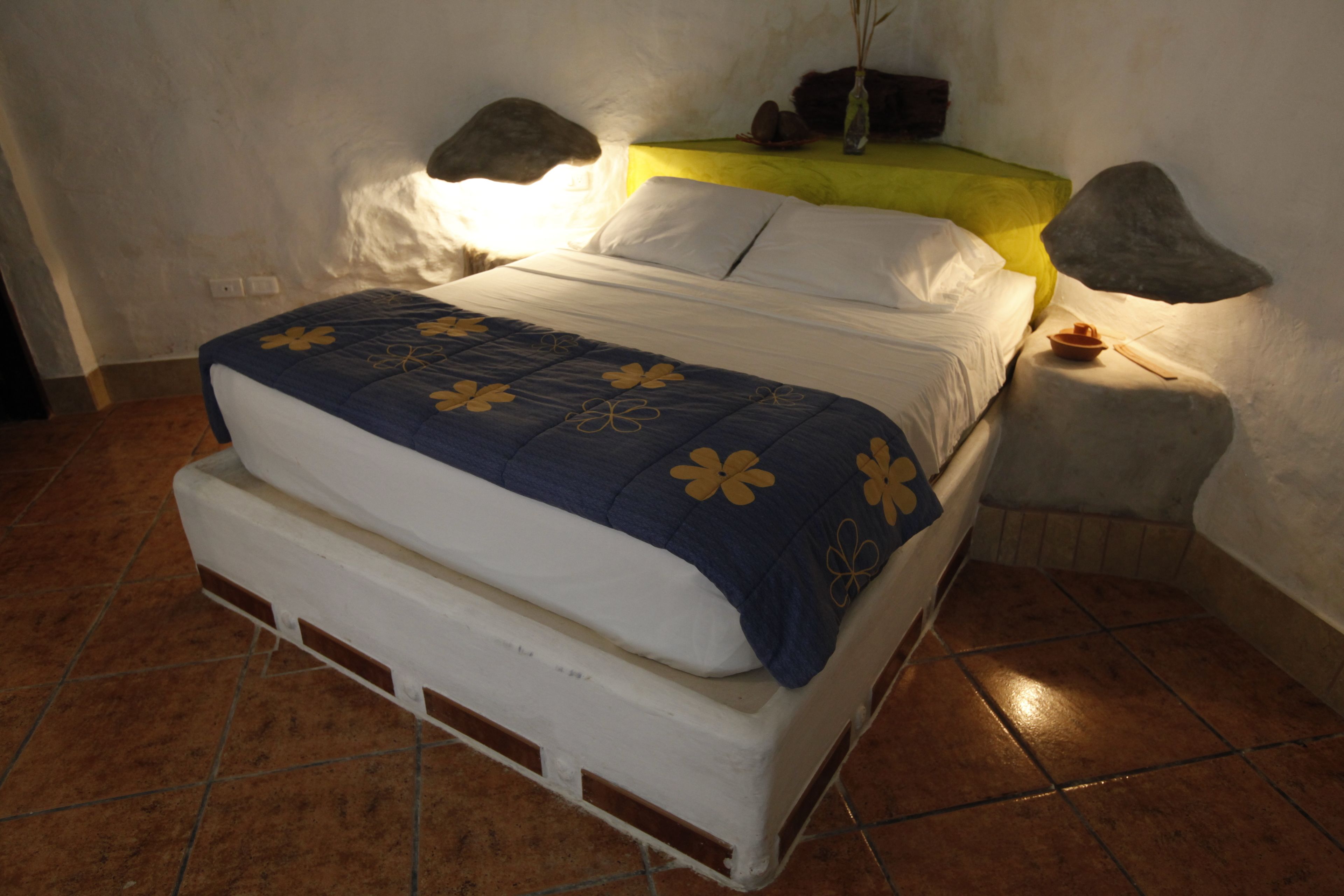 Una cama con una colcha azul y blanca, una mesita de noche con una lámpara, y una pequeña mesa con un tazón y un palito.