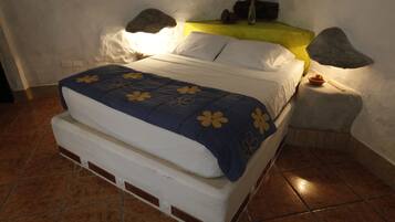 Una cama con una colcha azul y blanca, una mesita de noche con una lámpara, y una pequeña mesa con un tazón y un palito.