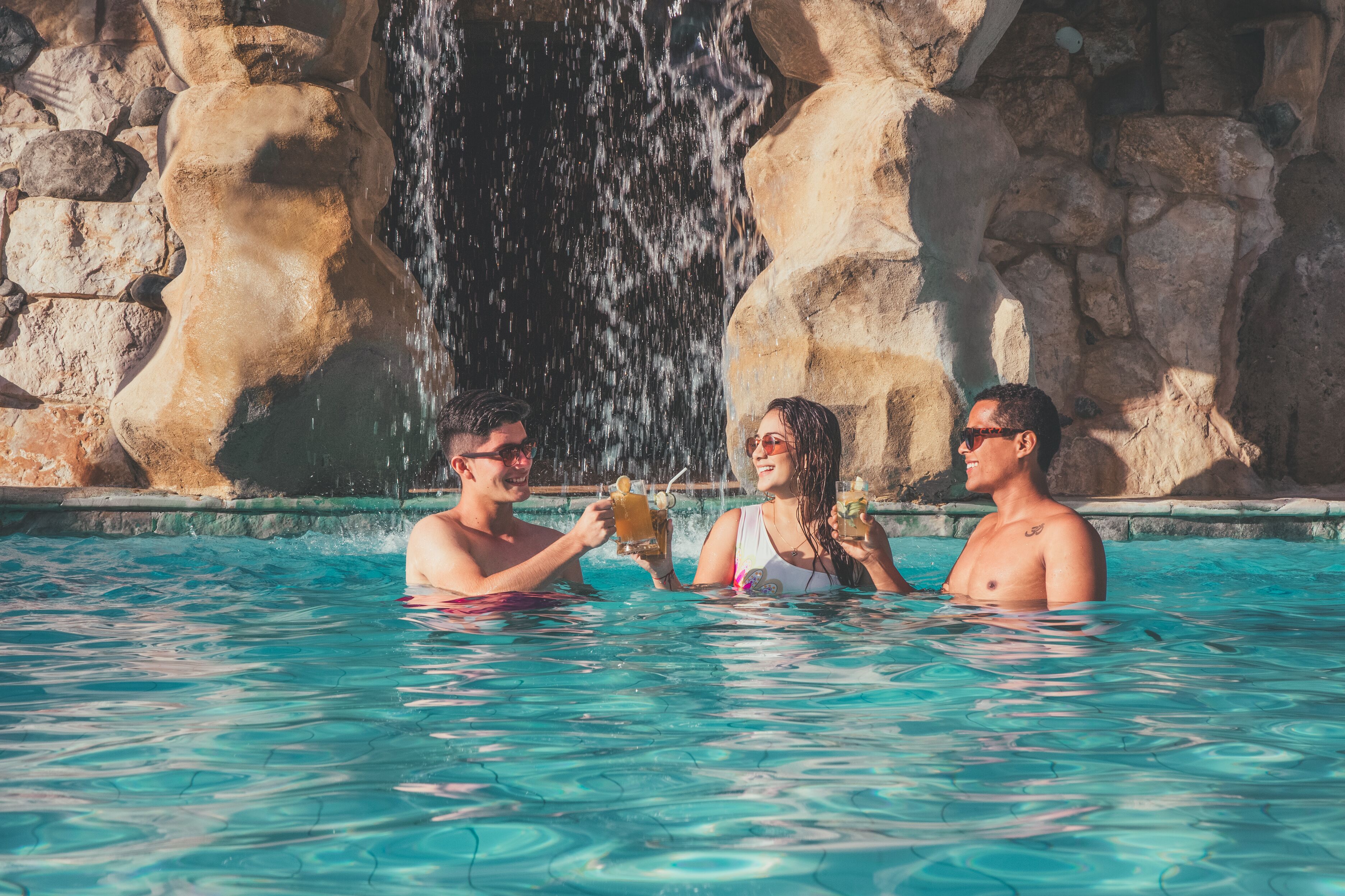 A pool with a waterfall backdrop, clear water, and people holding drinks.