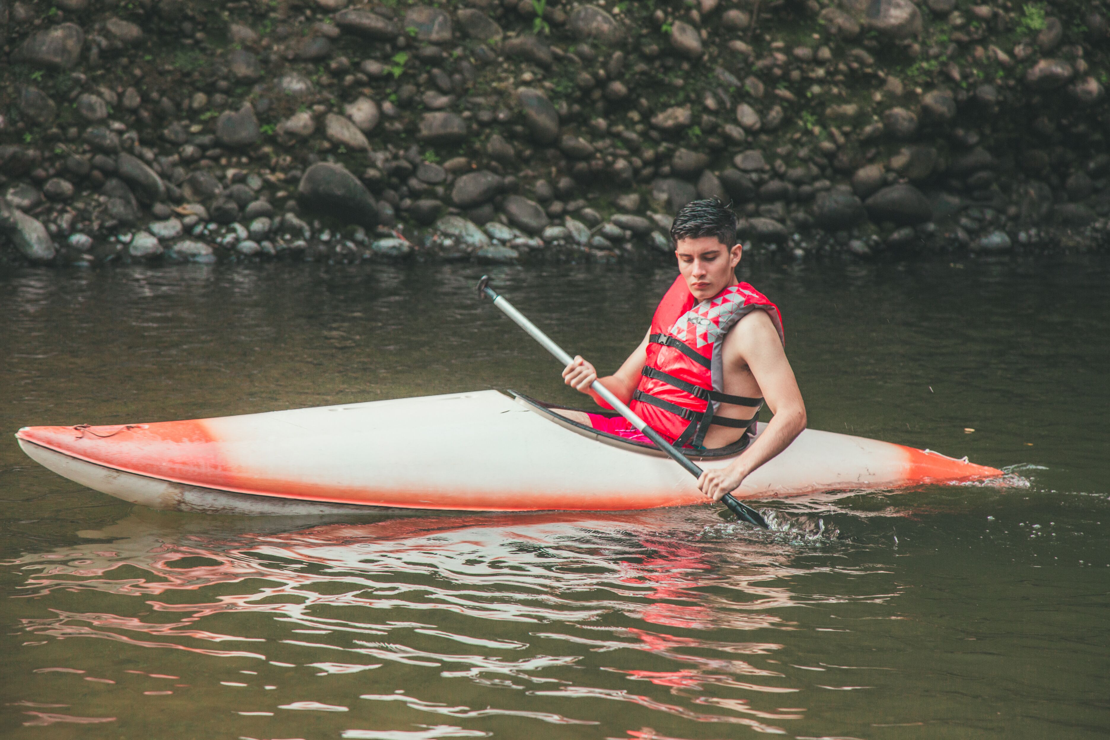 A person in a red life jacket paddling a white and orange kayak on calm water.