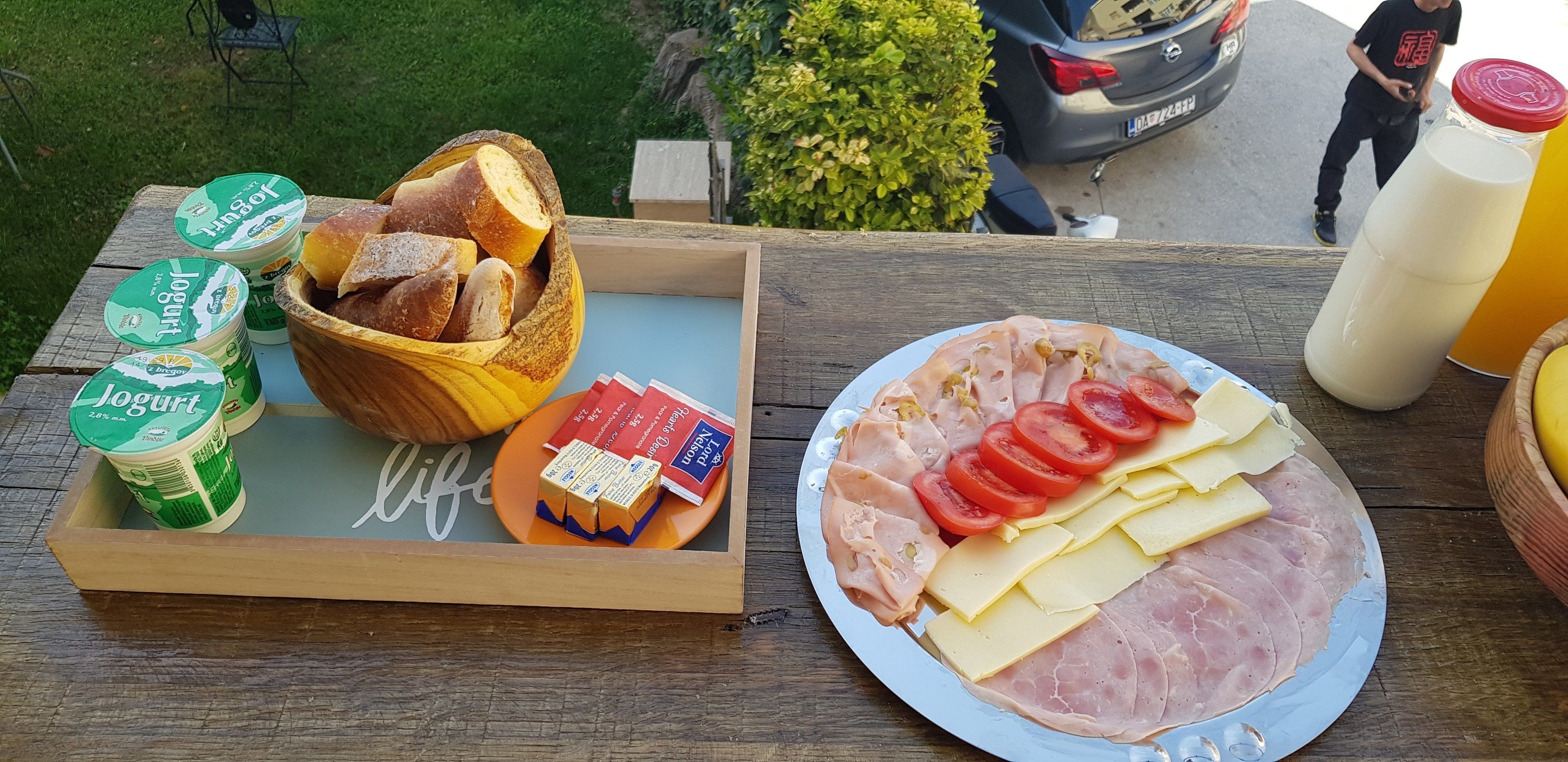 A picnic setup with bread, yogurt, cheese, ham, and tomatoes on a wooden table.