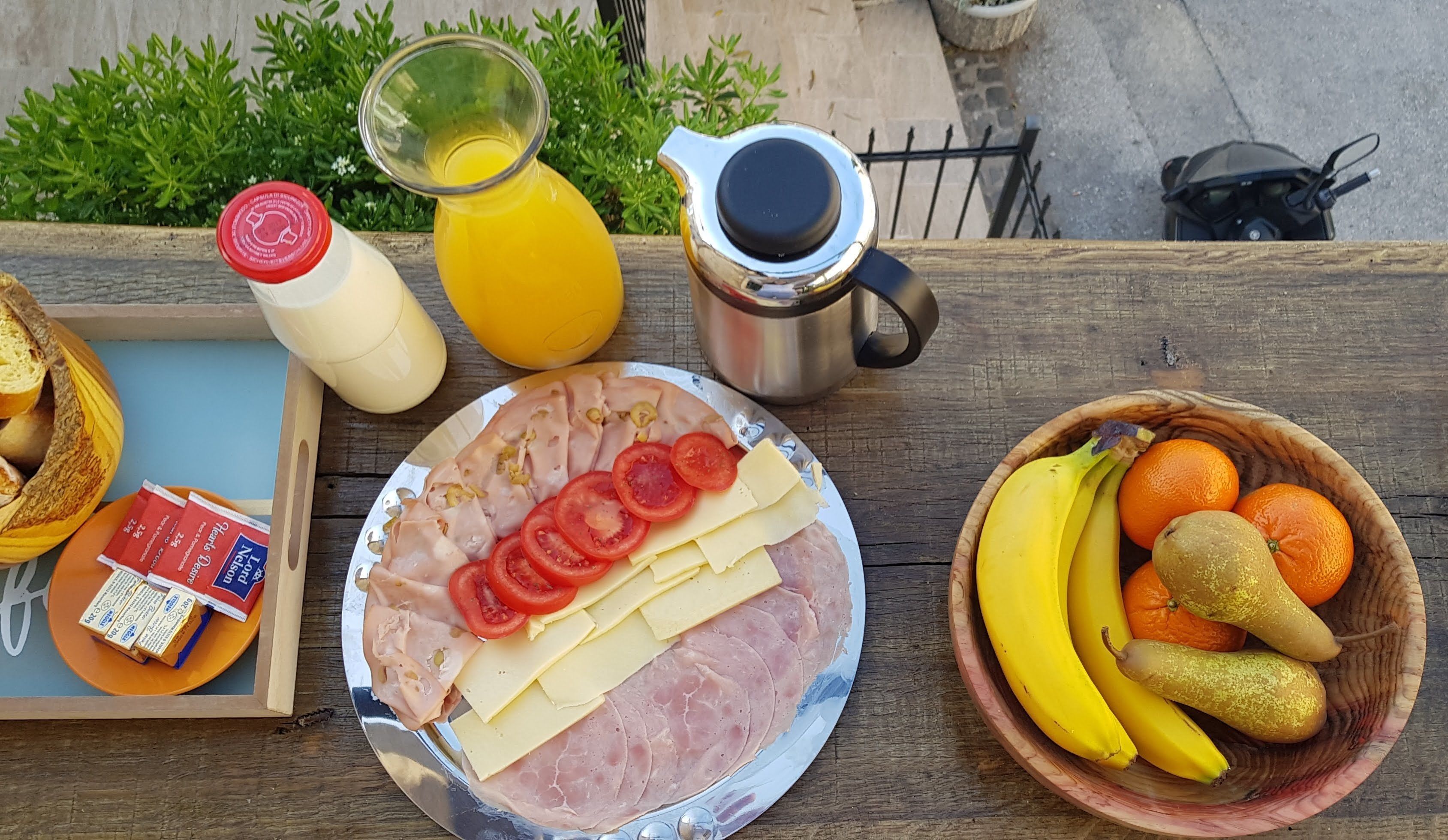 Una mesa de desayuno con jamón, queso, tomates, un bol de fruta, un vaso de jugo de naranja, una botella de leche y un termo.