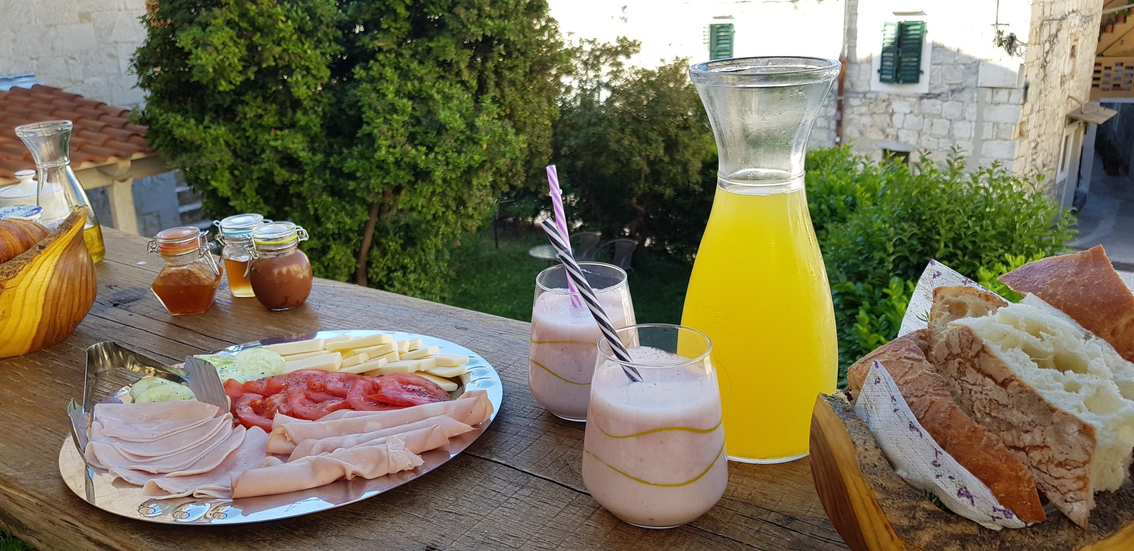 A picnic setup with a plate of sliced meats, vegetables, and bread, accompanied by drinks and condiments on an outdoor table.