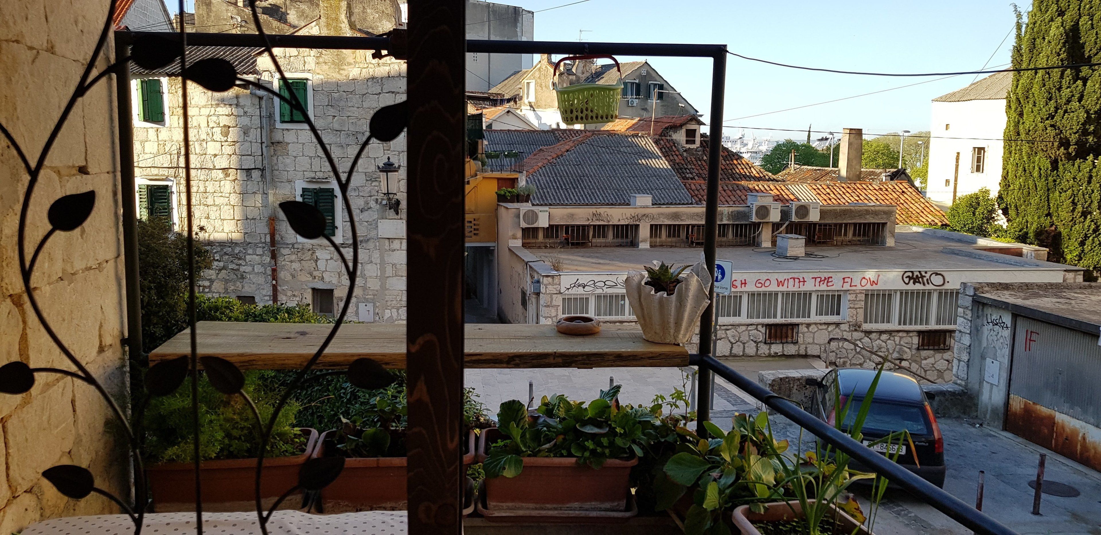 Un balcón con plantas en macetas, vista a una calle con edificios, y un letrero que dice "SH GO WITH THE FLOW".