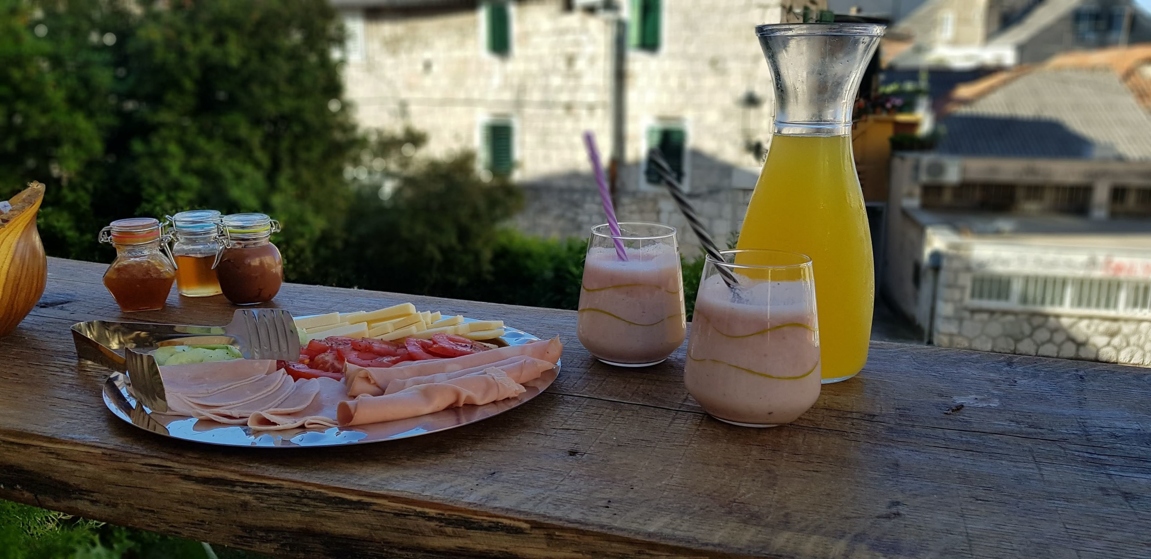 A picnic setup with a variety of food items and drinks on a wooden table, overlooking a building with green shutters.