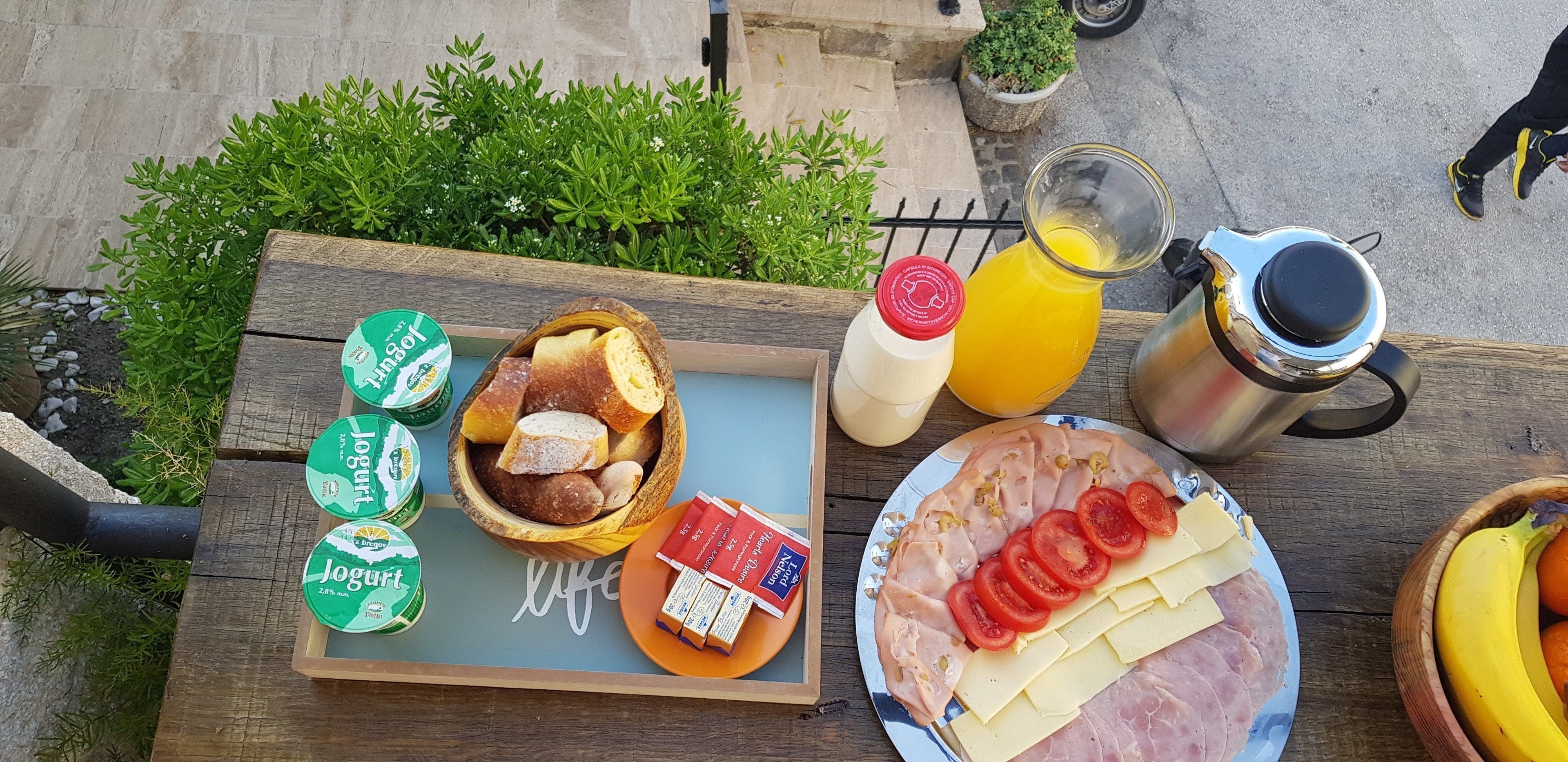 Una mesa de madera con un desayuno que incluye pan, yogur, queso, jamón, tomates y una banana.