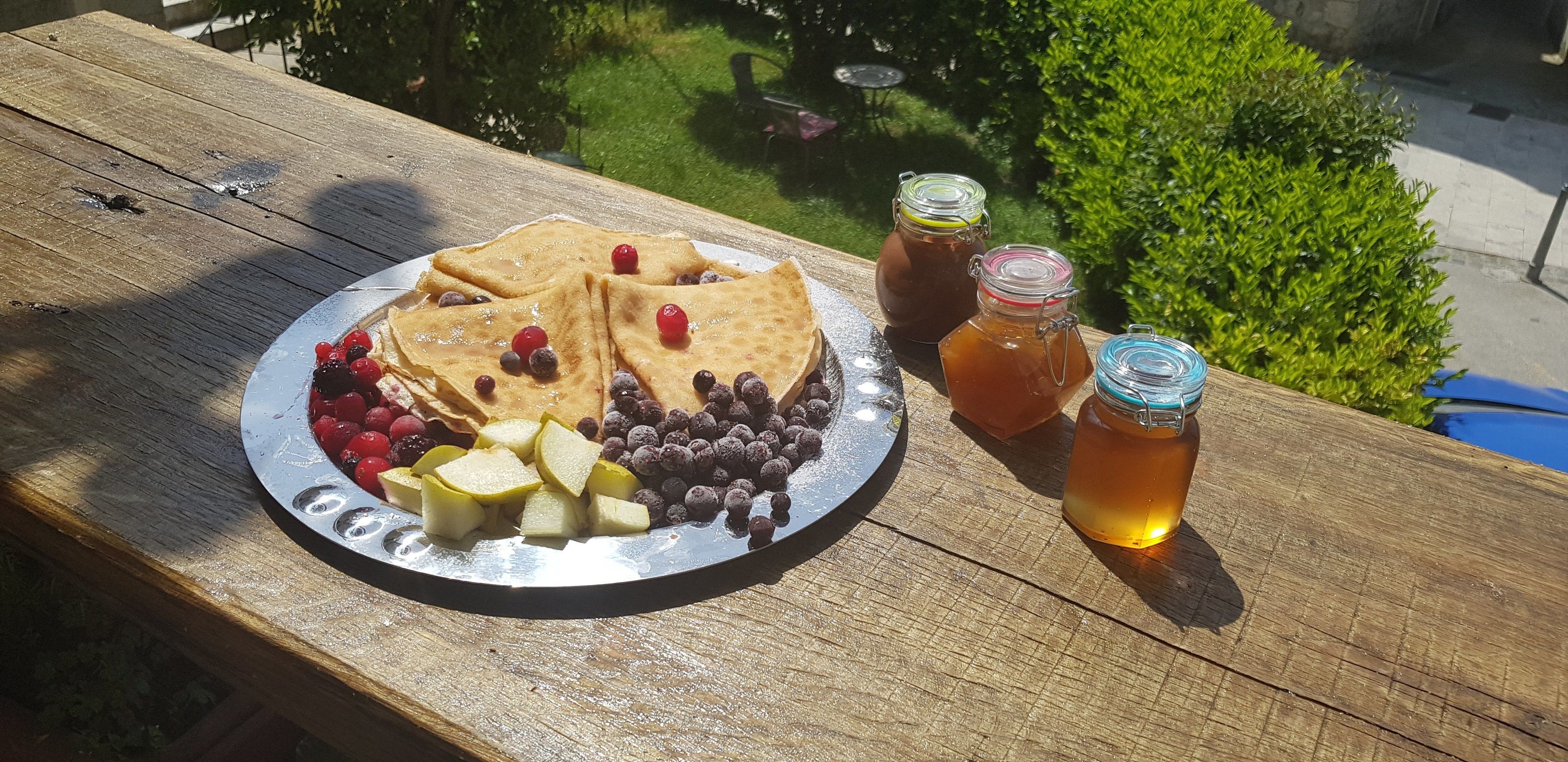 Una mesa de madera con una porción de panqueques, frutos rojos y manzanas, junto con dos tarros de miel.