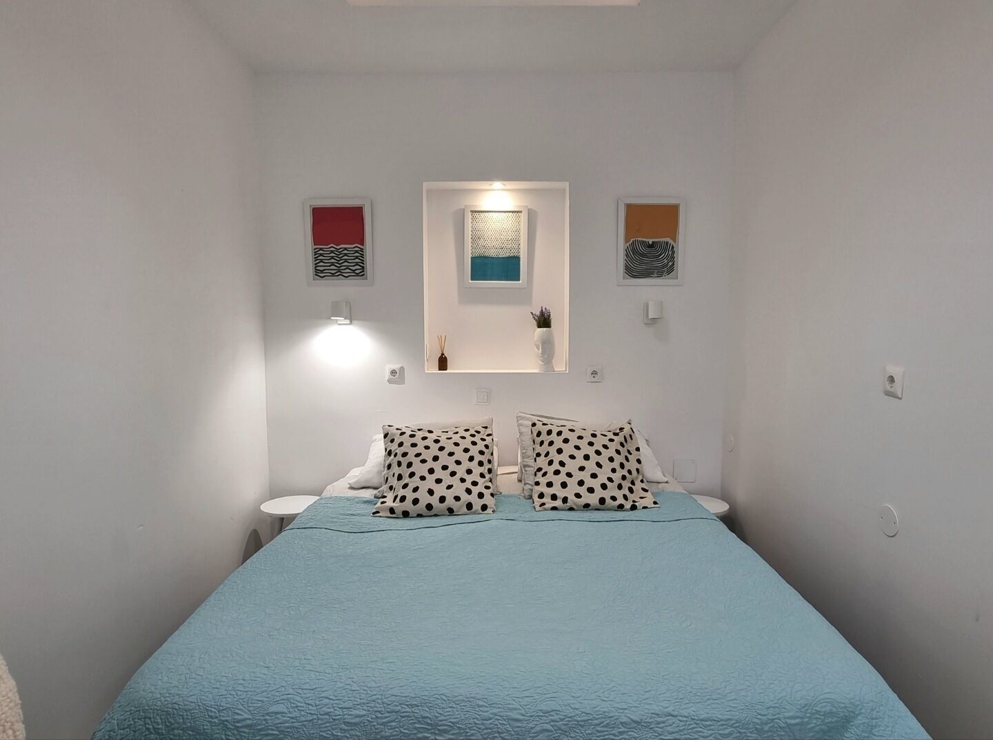 Piccola camera con un letto singolo, copriletto blu, pareti bianche e arredo essenziale, tra cui due lampade a parete e una piccola mensola con un vaso.