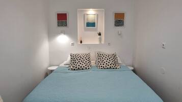 Piccola camera con un letto singolo, copriletto blu, pareti bianche e arredo essenziale, tra cui due lampade a parete e una piccola mensola con un vaso.