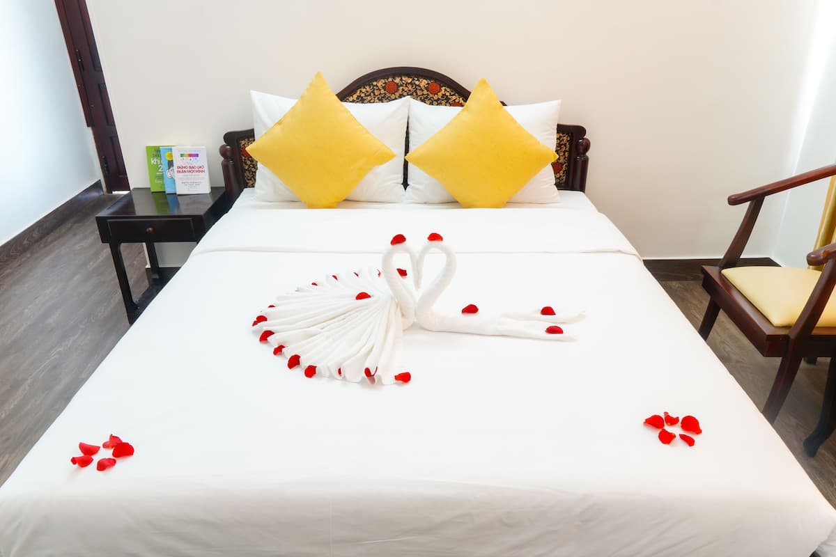 A neatly made bed with a swan and rose petals, a wooden nightstand with a book, and a chair with a cushion.