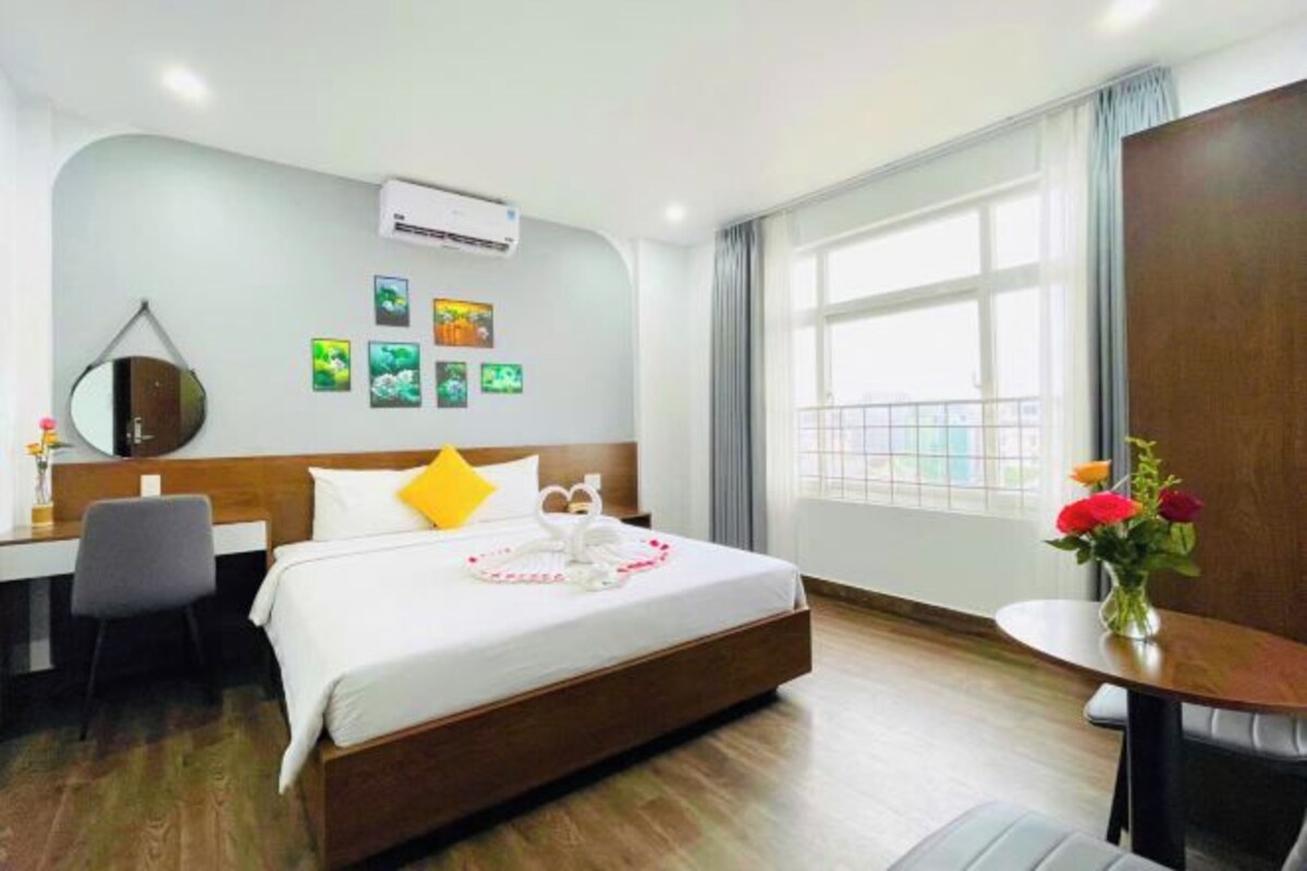 A hotel room with a bed, a desk, a chair, a small table with a vase of flowers, and a window with curtains.