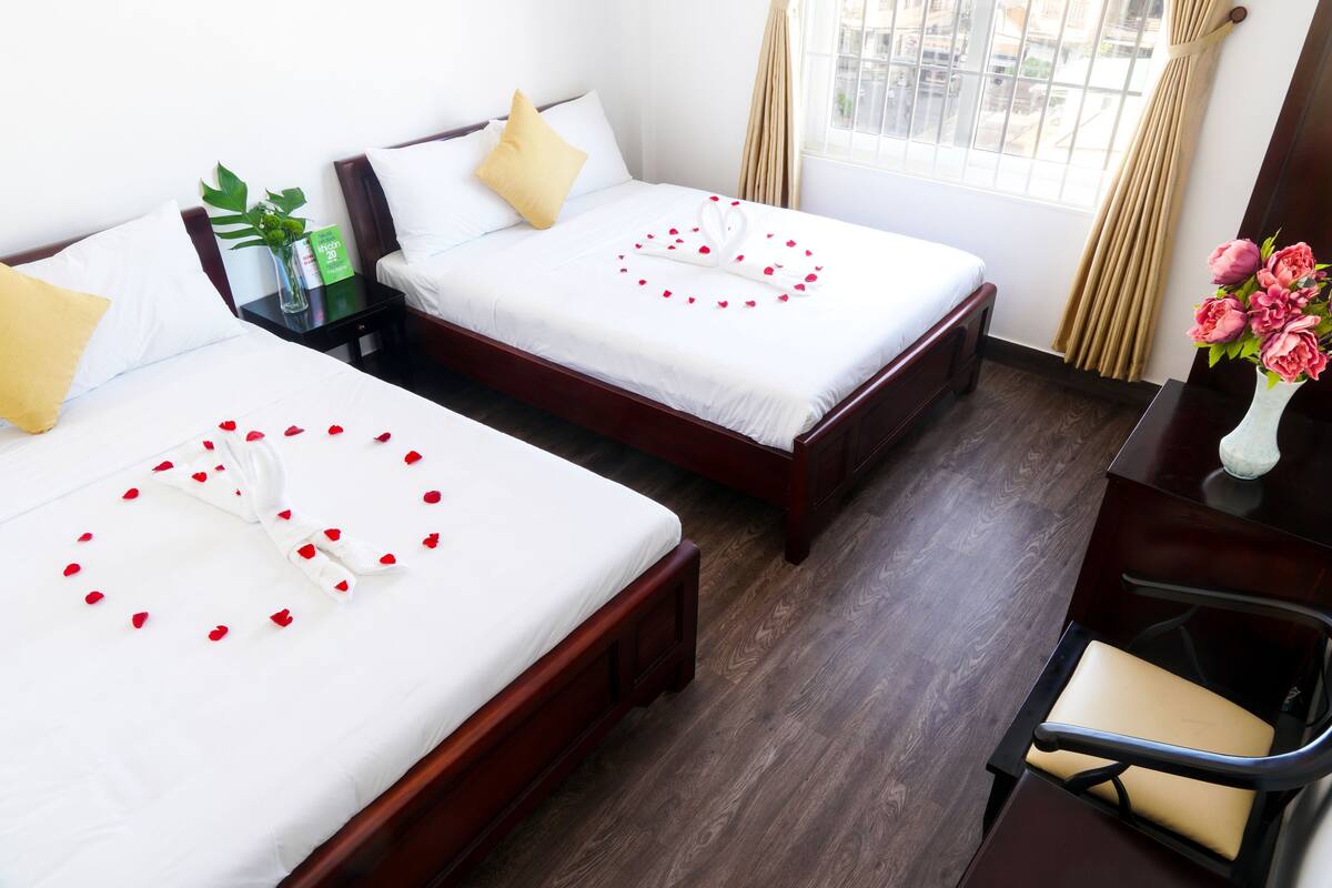 A hotel room with two beds, a desk, a chair, a vase with flowers, and a window with curtains.
