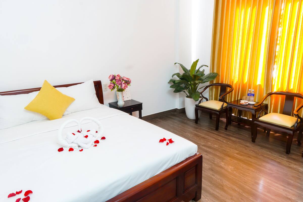 A hotel room with a wooden bed, a yellow pillow, a white bedspread, a bouquet of flowers, a potted plant, and two wooden chairs.