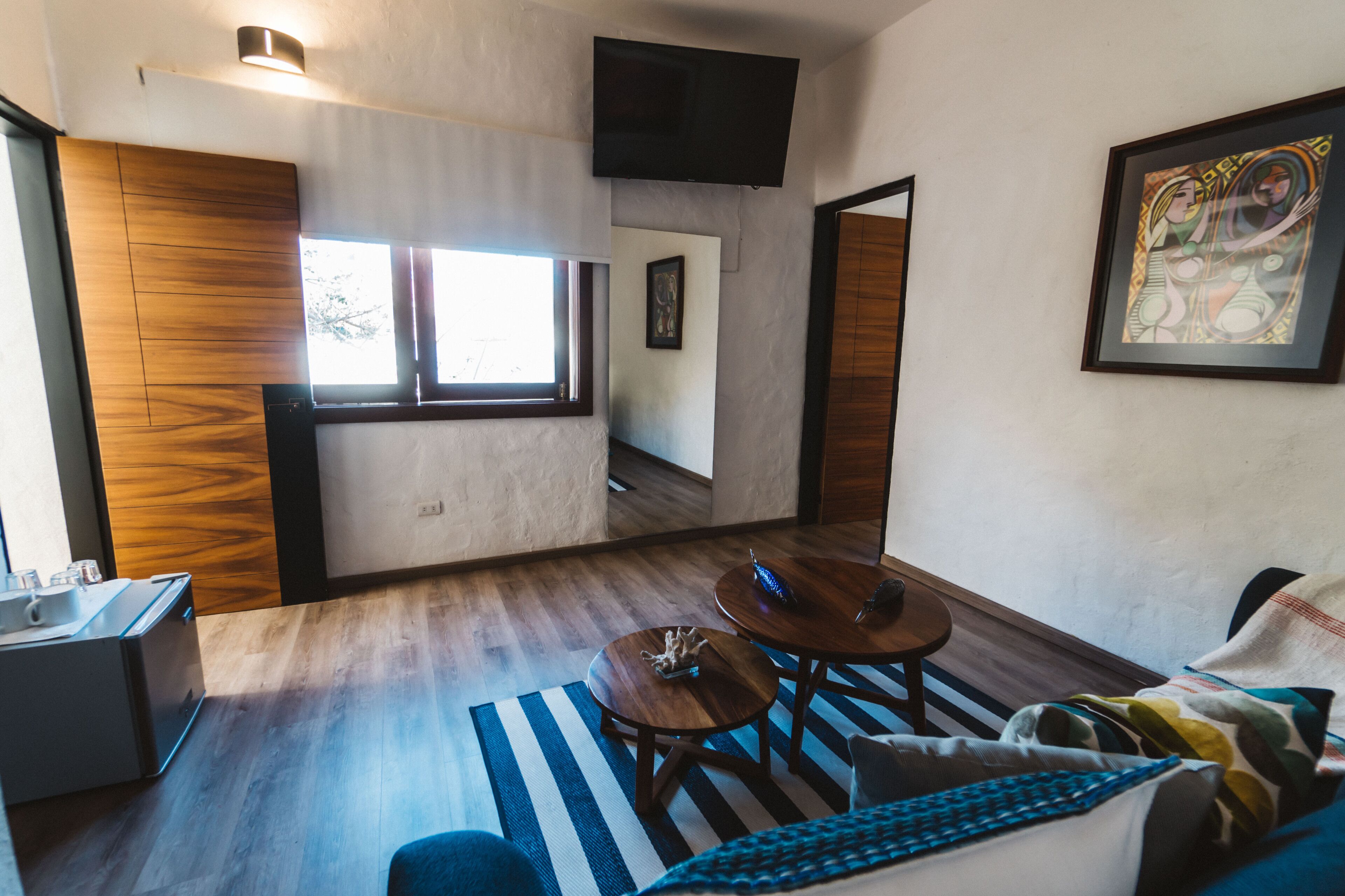 Un salon moderne avec un coin cuisine, un plancher en bois, un téléviseur à écran plat fixé au mur et une grande fenêtre qui laisse entrer la lumière naturelle.