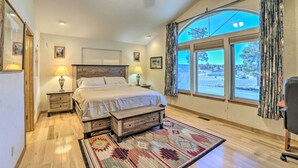 Une chambre avec un grand lit, deux tables de chevet avec des lampes, un banc en bois, un tapis à motifs et une grande fenêtre avec des rideaux.