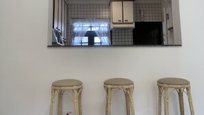 Eine Küche mit weißen Schränken, einem Fenster mit Spitzenvorhängen und einem eingebauten Herd. Vor der Küche stehen drei Korbstühle.