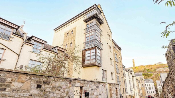 En smal brostensgade med gamle stenhuse, hvor et hjørnehus skiller sig ud med en balkon.