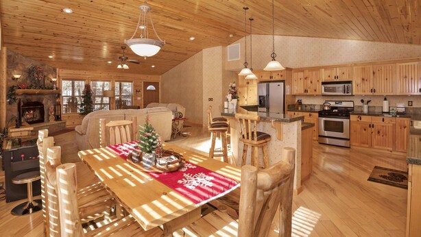 A spacious living area with a wooden ceiling, a fireplace, and a dining table with chairs. - Mountain Bliss by Destination Big Bear : Remarkable Single Story Log Style Home in Pinewood Estates! Outdoor Hot Tub! Arcade! (Big Bear City)