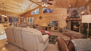 A cozy log cabin interior with a fireplace, a large sofa, and a television. - Mountain Bliss by Destination Big Bear : Remarkable Single Story Log Style Home in Pinewood Estates! Outdoor Hot Tub! Arcade! (Big Bear City)