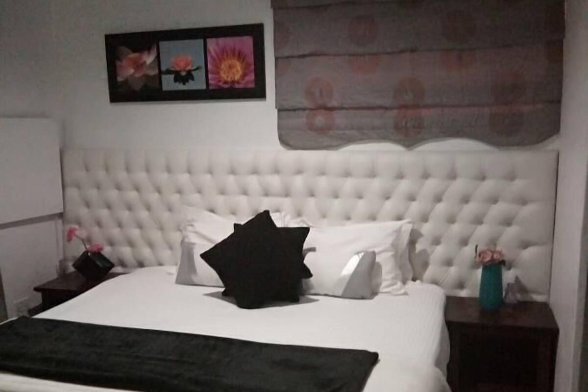 Um quarto com cabeceira estofada, uma cama grande com roupa de cama branca, um cobertor preto e uma mesa de cabeceira com um vaso de flores.