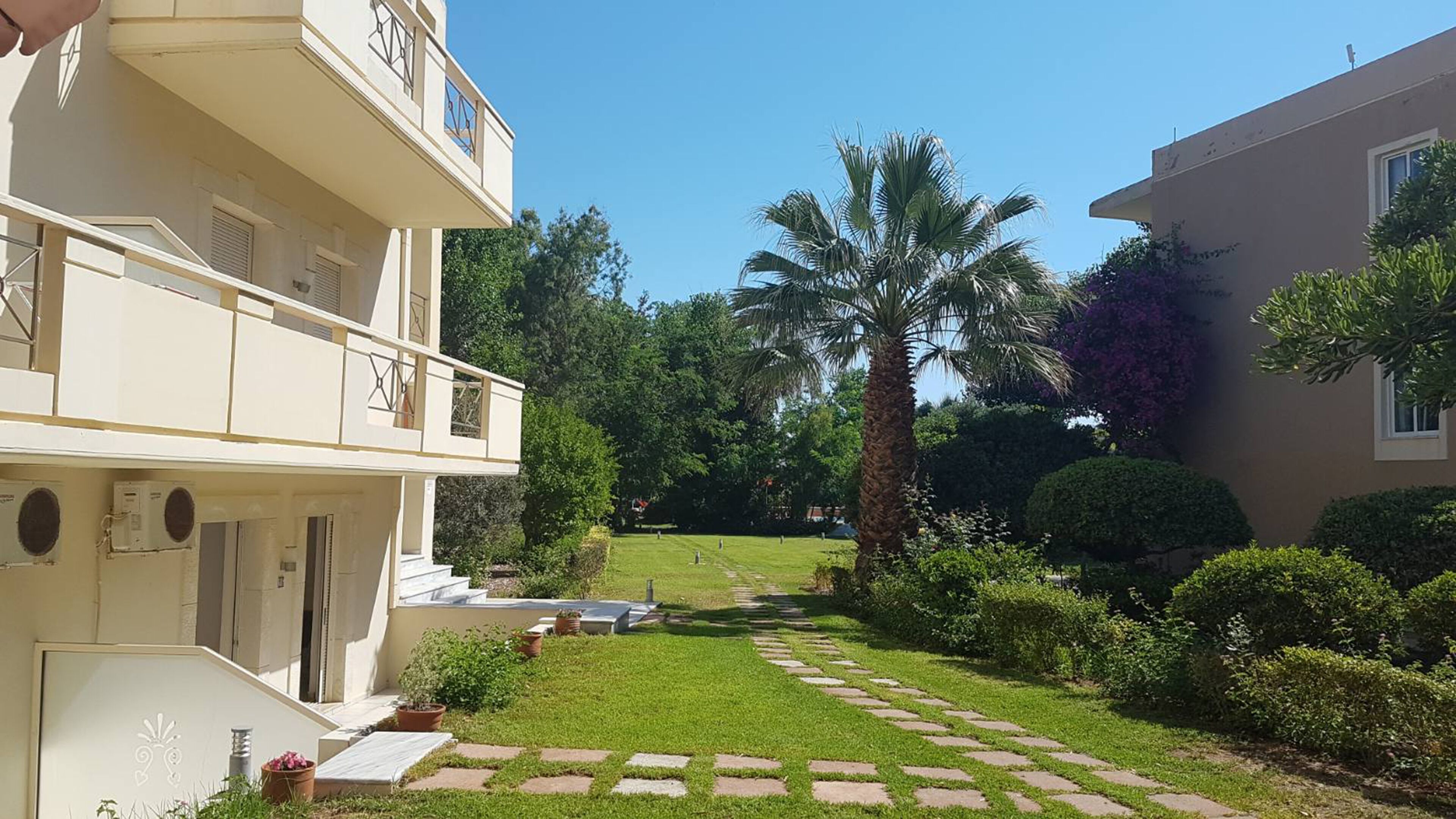 Un bâtiment à deux étages doté d’un balcon, d’un jardin bien entretenu avec un palmier, et un ciel dégagé.