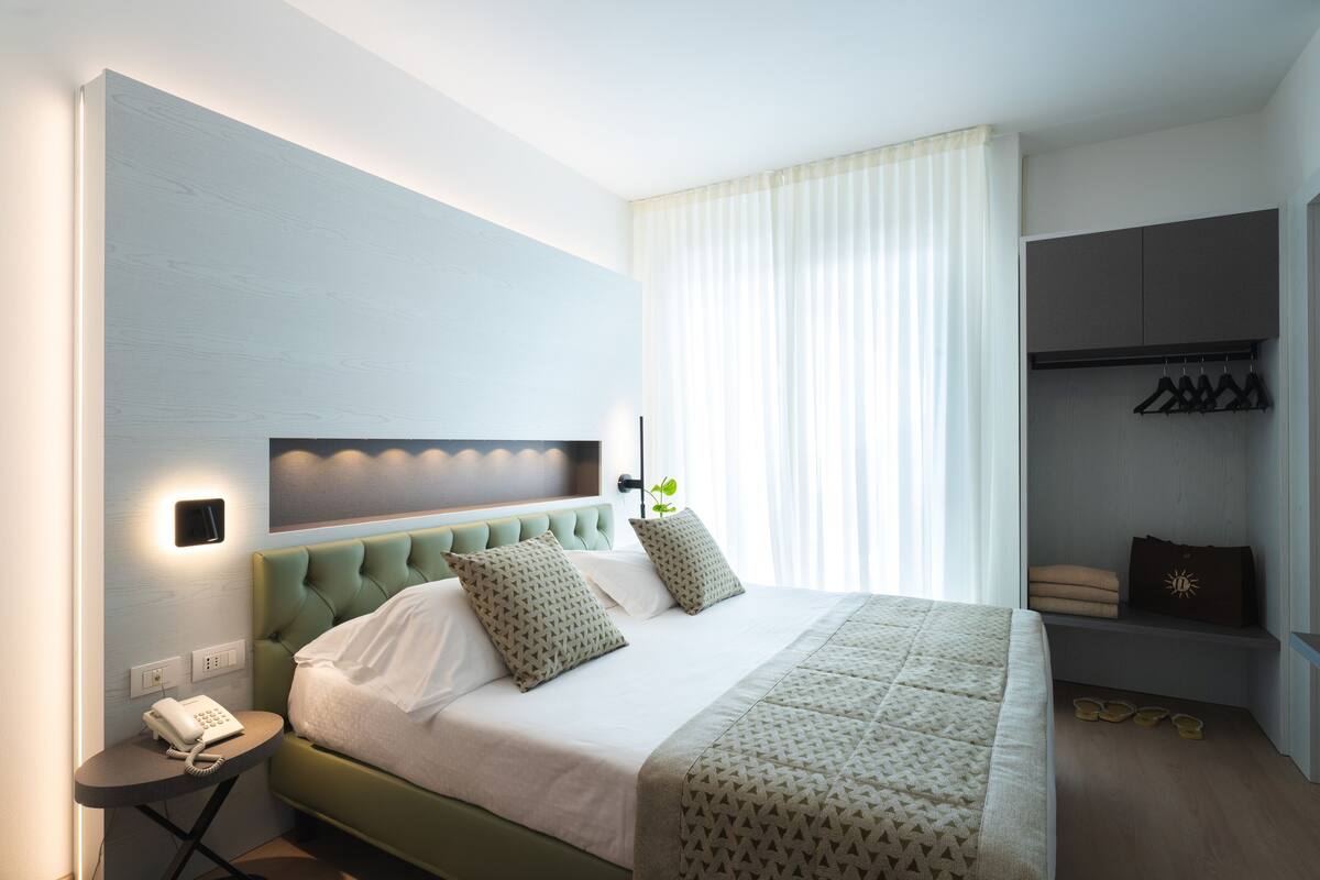 A modern hotel room with a green upholstered headboard, a bedside telephone, a neatly made bed with patterned pillows, and a small seating area with a bench and hanging clothes.